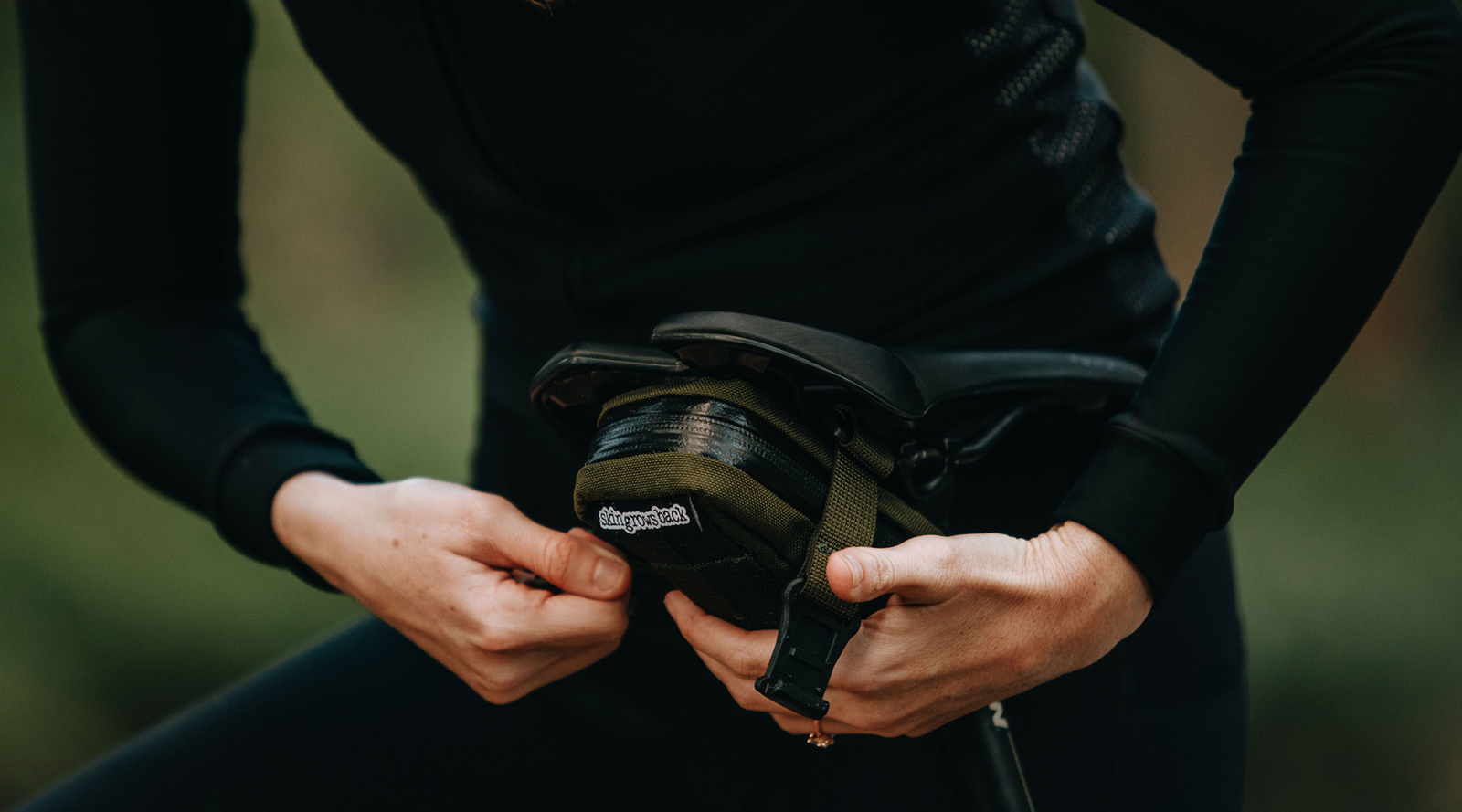 Skingrowsback Plan B Saddle Bag in olive — woman in black attaching it to her bike saddle